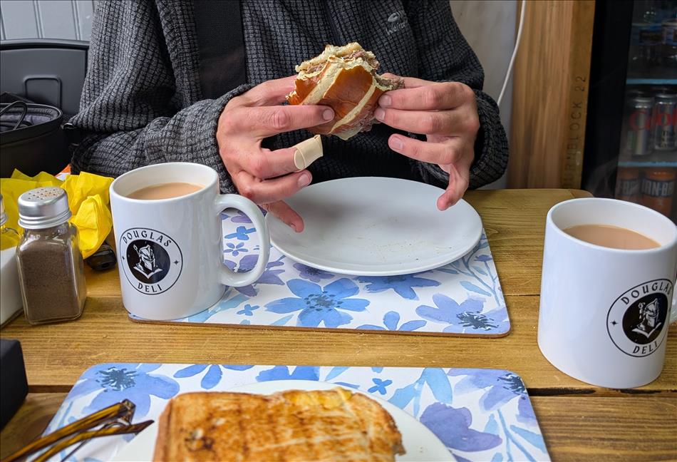 Ren holds his burger, we see a plaster on his middle finger. The table has tea and Sharon's butty too