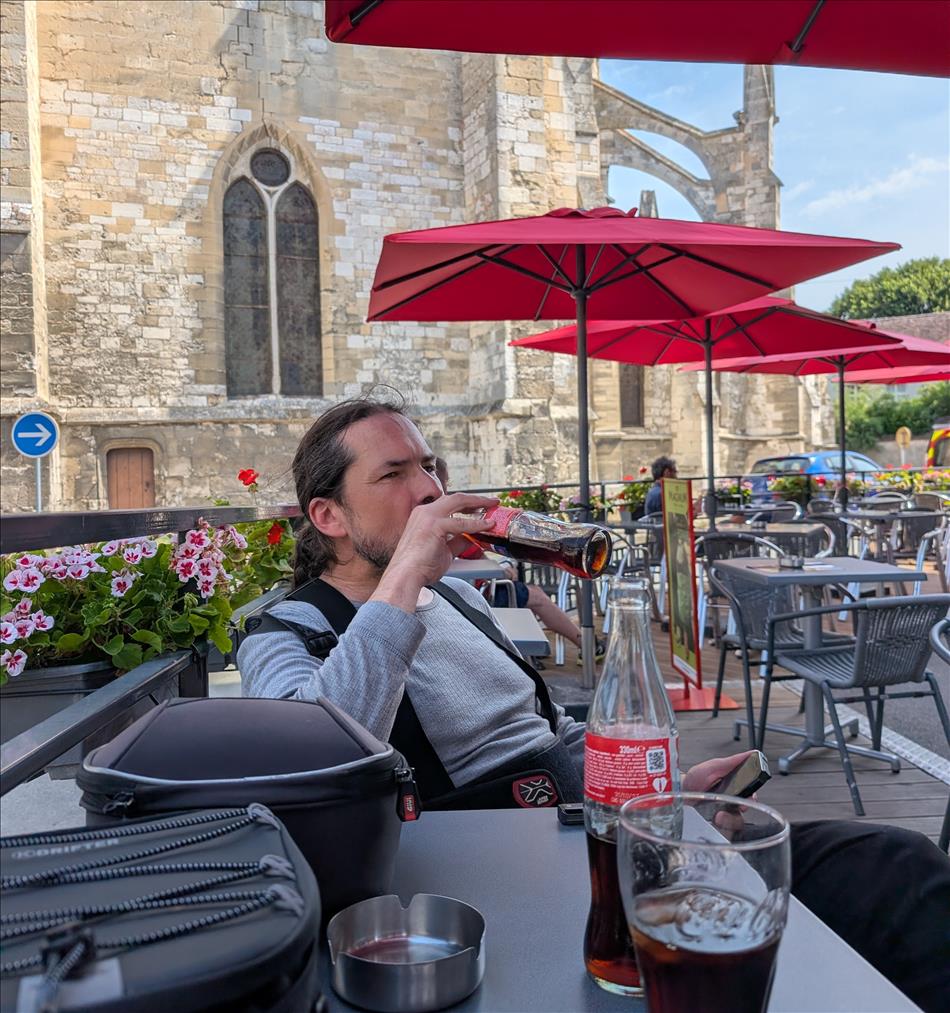 Ren drinks from a cola bottle, sat at the table under parasols at the cafe